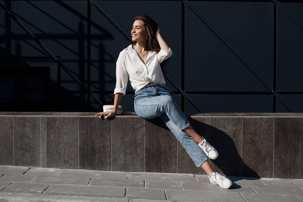 A brunette woman with long hair in fashionable in blue jeans and a white shirt posing outdoors. Fashion photo concept. Woman near business center. Portrait of a brunette woman.Beautiful female business center worker near her office - Photo, Image
