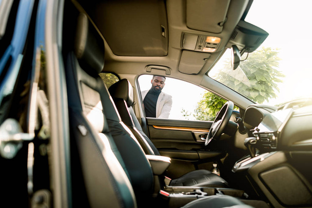 Nuori afrikkalainen liikemies avaa uuden autonsa oven. Sivunäkymä, aurinkoinen päivä
 - Valokuva, kuva