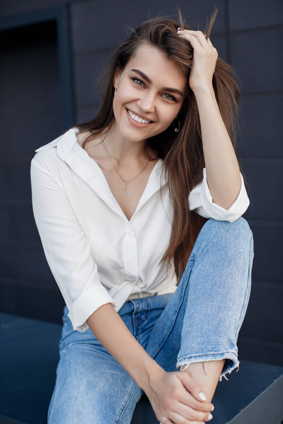 A brunette woman with long hair in fashionable in blue jeans and a white shirt posing outdoors. Fashion photo concept. Woman near business center. Portrait of a brunette woman.Beautiful female business center worker near her office - Foto, Bild