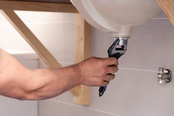 Zbliżenie męskich hydraulików ręcznie naprawić zlewozmywak w łazience z płytkami ściennymi. Profesjonalny serwis hydrauliczny, instalacja rur wodnych. Klucz w odpływie ścieków montowany przez człowieka - Zdjęcie, obraz