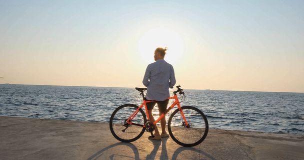 Genç ve yakışıklı bir erkek, günbatımına ve denize karşı sabah sahilinde renkli bisiklete biniyor. - Fotoğraf, Görsel