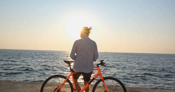 Genç ve yakışıklı bir erkek, günbatımına ve denize karşı sabah sahilinde renkli bisiklete biniyor. - Fotoğraf, Görsel