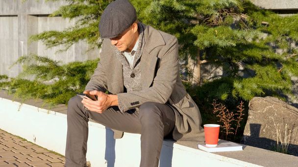 Young stylish male with cellphone and coffee in park - 写真・画像