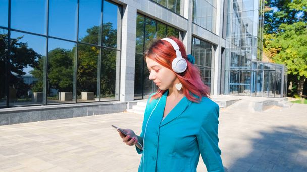 Young happy modern businesswoman with pink hair and colorful suit walk outdoors and listen to music with headphones - Valokuva, kuva