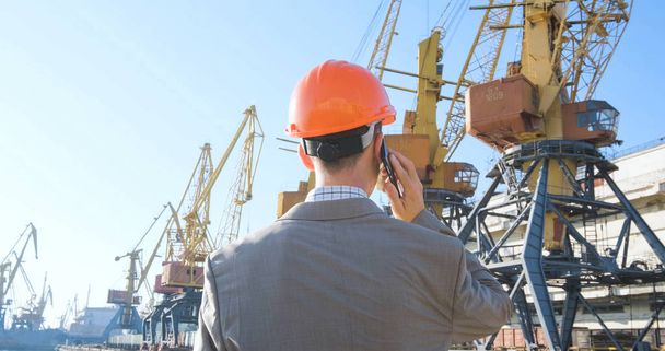 Young male worker of sea harbor in helmet, cargo manager in suit and halmet works outdoor , cranes and sea background - Photo, Image