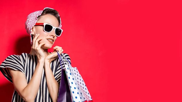 Happy shopping woman with shopping bags over bright red background wearing hat and sunglasses - Photo, Image