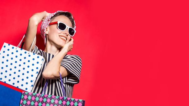 Happy shopping woman with shopping bags over bright red background wearing hat and sunglasses - Фото, изображение