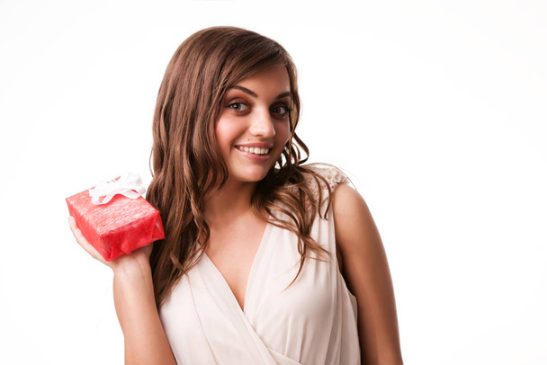 Young beautiful positive brunette woman in beige dress standing, holding small red gift box in hand and smiling - Фото, изображение