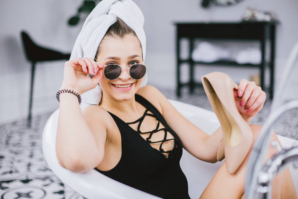 Fashion shot of shopping, young caucasian woman in sunglasses and towel on head holding high heels shoes in hand, lying in bathroom at hotel near window - Фото, зображення