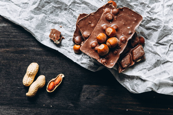 Melkchocolade met noten: pinda 's, hazelnoten op wit perkamentpapier met notenkraker en op een donkere houten tafel. Selectieve focus - Foto, afbeelding