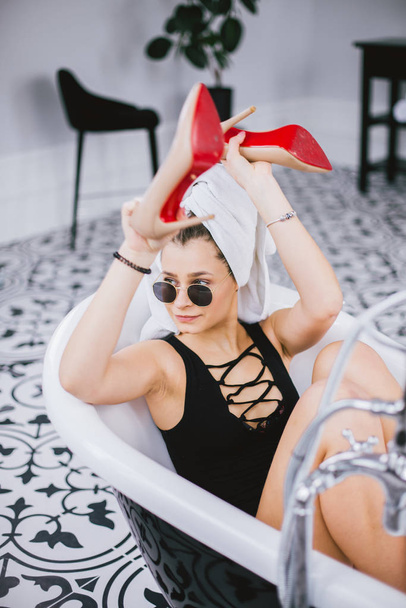Fashion shot of shopping, young caucasian woman in sunglasses and towel on head holding high heels shoes in hand, lying in bathroom at hotel near window - Photo, Image