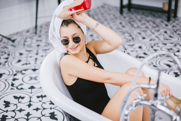 Moda tiro de compras, joven mujer caucásica en gafas de sol y toalla en la cabeza con zapatos de tacón alto en la mano, acostado en el baño en el hotel cerca de la ventana
 - Foto, Imagen