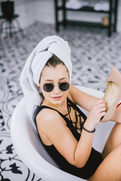 Prise de vue de mode de shopping, jeune femme caucasienne en lunettes de soleil et serviette sur la tête tenant des chaussures à talons hauts à la main, couché dans la salle de bain à l'hôtel près de la fenêtre
 - Photo, image