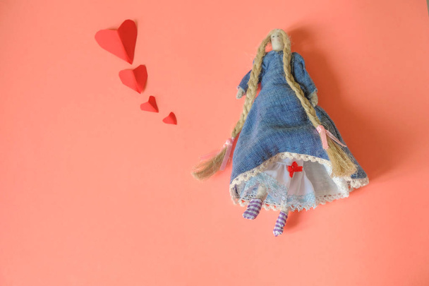 Grappig klein zakpop gemaakt van stof in vliegende rokken en met lange linnen vlechten op een koraal achtergrond. De pop bekent in liefde. Kopieerruimte. - Foto, afbeelding