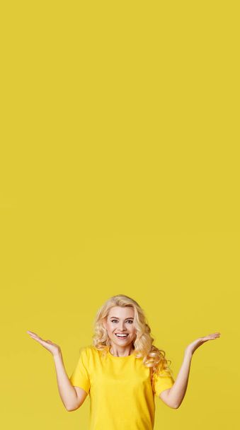 junge glückliche Frau zeigt Hände nach oben, breitet seine Hände zur Seite aus. oben ist ein leerer Platz für Text und Beschriftung auf gelbem Hintergrund. Senkrechtschuss - Foto, Bild