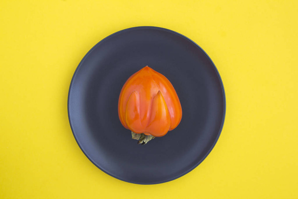 Ugly food. Ugly persimmon on the black plate  in the center of the yellow background. Top view. Copy space. - Photo, Image