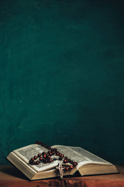 Otwórz Pismo Święte i krucyfiks koralików na czerwonym starym drewnianym stole. Piękne zielone tło ściany. - Zdjęcie, obraz