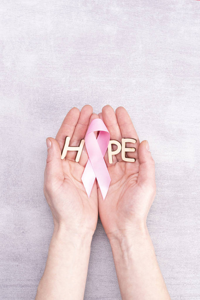 egészségügy és orvostudomány koncepció - női kéz kezében rózsaszín mellrák tudatosság szalag és betűk a szó Hope szürke háttér - Fotó, kép