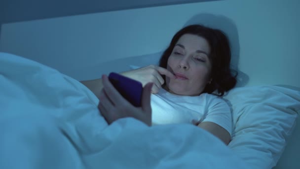 Woman using social media on smartphone at night, smiling and flirting online - Video