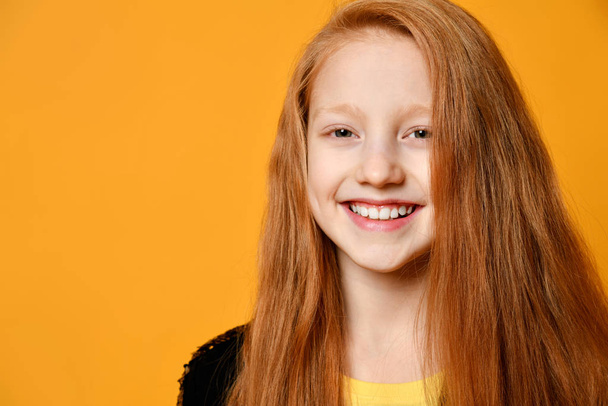 Red-haired teenage kid in black jacket. She is smiling and looking at you, posing against orange studio background. Close up - Zdjęcie, obraz