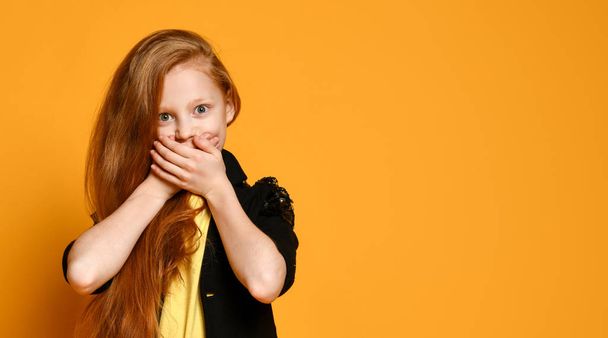 Teenage girl in black jacket , yellow t-shirt. She is shocked, covered her mouth, posing on orange background. Close up - Fotó, kép