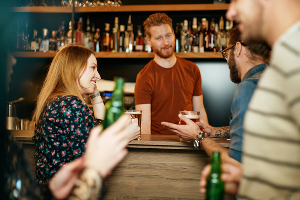 Un pequeño grupo de mejores amigos de pie en un pub, beber cerveza, charlar y divertirse. Concéntrate en el camarero limpiando vasos. Vida nocturna
. - Foto, imagen