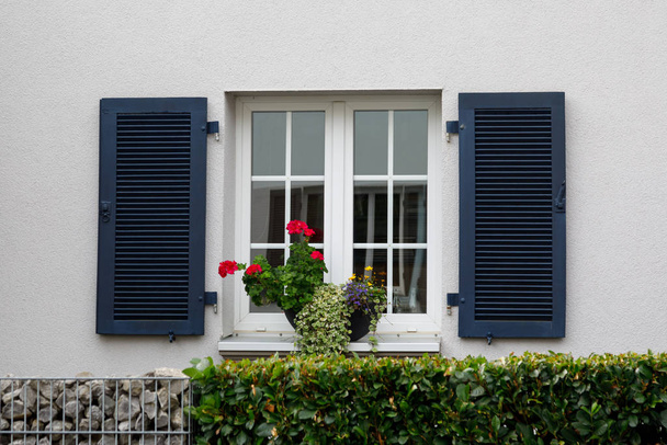 Egy ablak fehér kerettel, sötétkék redőnyökkel és virágokkal az ablakpárkányon. Egy hagyományos európai vidéki ház homlokzata. - Fotó, kép