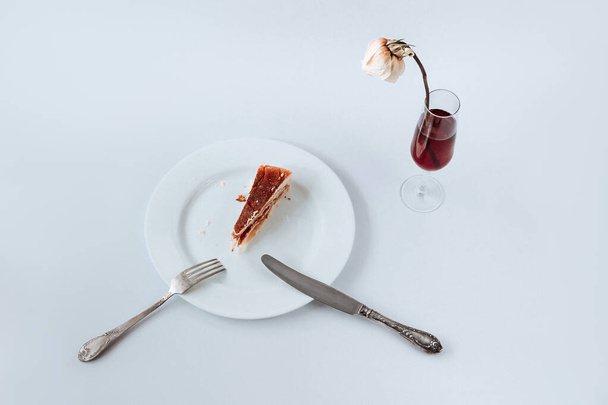 Witte plaat met een stuk taart erop, bestek en glas champagne met vervaagde roos op een witte achtergrond - Foto, afbeelding