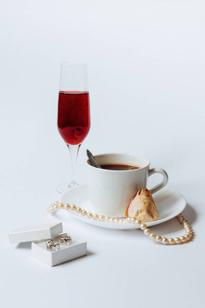 ein Stillleben mit einer weißen Keramiktasse Kaffee, einer Rosenknospe, einer Korallenkette, einer kleinen weißen Schachtel mit den silbernen Ohrringen und einem Glas Champagner auf weißem Hintergrund - Foto, Bild
