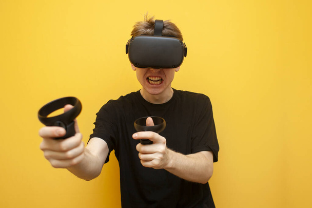 парень в очках VR на жёлтом фоне играет виртуального шутера, эмоциональный геймер стреляет в игру джойстиками и криками
 - Фото, изображение