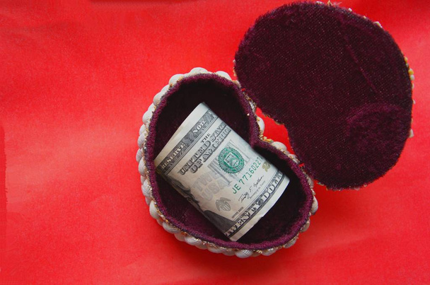 Деньги лежат в декоративной коробке на красном фоне в виде сердца
 - Фото, изображение