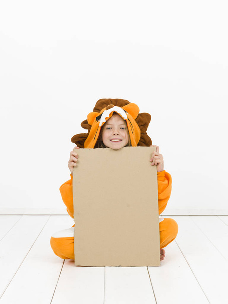 pretty blonde girl in cozy lion costume posing with cardboard blank banner in front of white wall - Фото, изображение