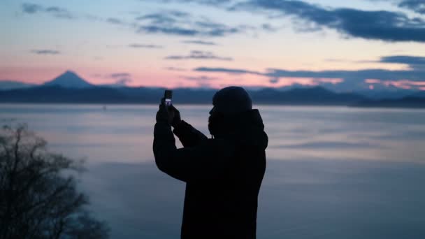 Man Takes a Picture with His Smartphone in a Snowy Wild Landscape at Sunset. Backview of one lonely adult guy spending time by the going and look around on the beauty pictures of wild seaside area. - Záběry, video