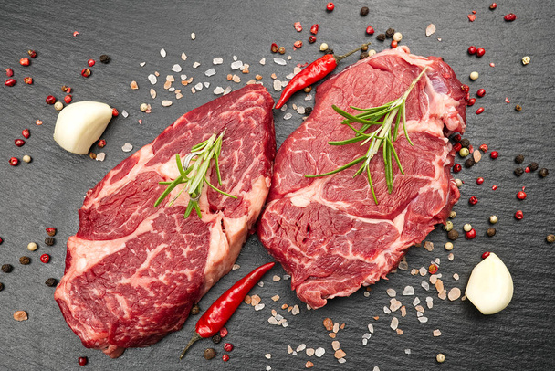 Raw rib eye steak with spices and vegetables. Ingredients for restaurant meal. Fresh meat, salt, rosemary, thyme, chilli, cherry tomatoes, garlic on black stone. Food background. - 写真・画像