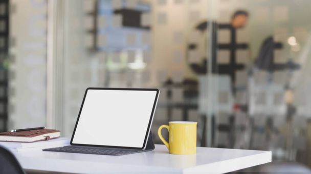 Vista cercana del lugar de trabajo con tableta de pantalla en blanco, suministros de oficina y taza de café en la mesa blanca con sala de oficina borrosa
  - Foto, Imagen