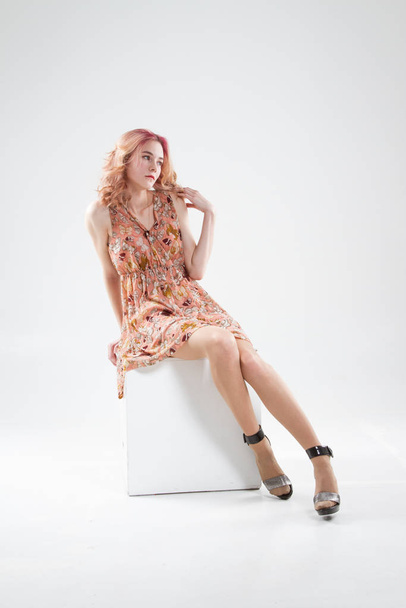 young blonde in a short dress sits on a cube. Studio portrait on a white background - Foto, imagen