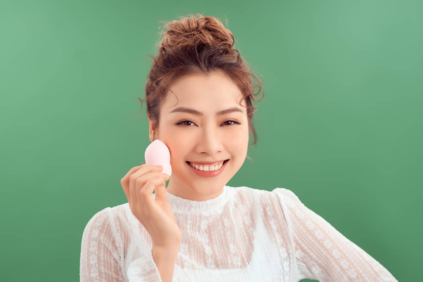 Mujer asiática riendo y sosteniendo esponja facial aislada sobre fondo verde
 - Foto, imagen