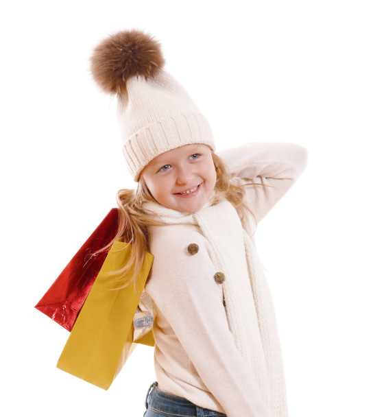 A little girl in a knitted hat with a pompon, scarf and sweater, is holding multicolored paper bags. Isolated on white background - Fotó, kép
