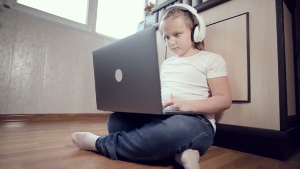 A smart little girl of seven years old in white headphones with a laptop in her hands is pushing on the floor in her room. The young generation on the Internet and IT technology - Materiał filmowy, wideo