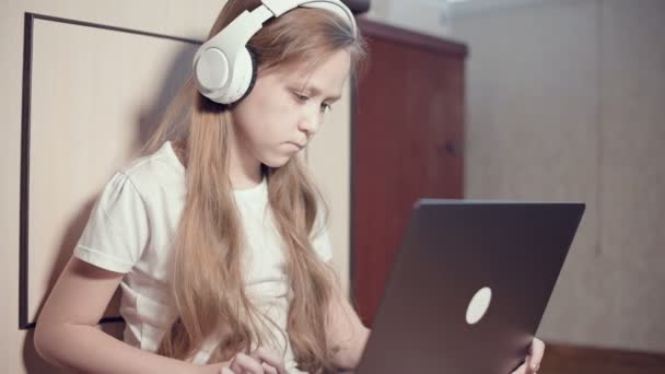 A smart little girl of seven years old in white headphones with a laptop in her hands is pushing on the floor in her room. The young generation on the Internet and IT technology - Filmmaterial, Video