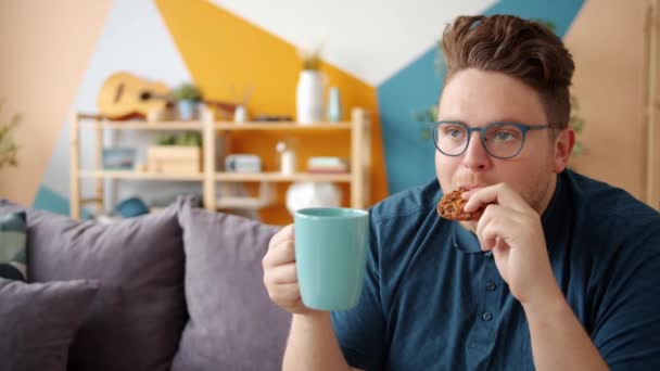 Portrait of cheerful man drinking coffee and eating biscuit alone in apartment - Filmati, video