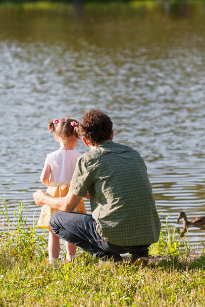 Πατέρας και κόρη κοιτάζουν τη λίμνη. Μια άγρια πάπια κολυμπάει μέσα στο νερό. Πίσω όψη. - Φωτογραφία, εικόνα