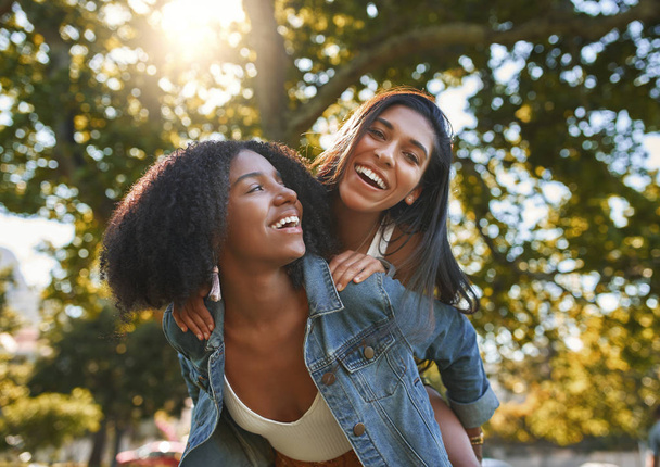 Portrait of a smiling african american young woman carrying her best friend on her back giving piggyback ride in park on a sunny day - friends laughing and having fun outdoors - Zdjęcie, obraz