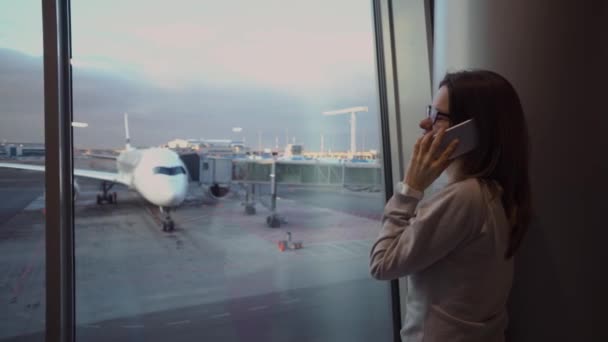 Νεαρό, όμορφο κορίτσι που μιλάει στο τηλέφωνο στο τερματικό του αεροδρομίου με φόντο ένα αεροσκάφος - Πλάνα, βίντεο