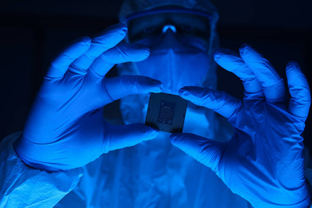 A man in a protective suit holds a microchip in his hands. Research lab worker wearing gloves and safety glasses. - Foto, Imagen