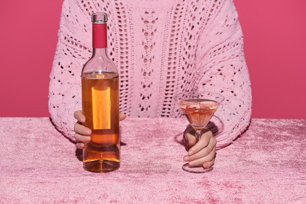 cropped view of woman holding bottle of alcohol drink and glass at velour pink table isolated on pink, girlish concept - Photo, Image
