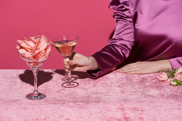 cropped view of woman holding glass of rose wine near glass with rose petals on velour cloth isolated on pink, girlish concept  - Foto, Bild