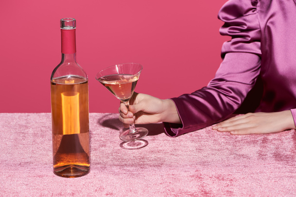 cropped view of woman holding glass of rose wine near bottle on velour cloth isolated on pink, girlish concept  - Fotografie, Obrázek