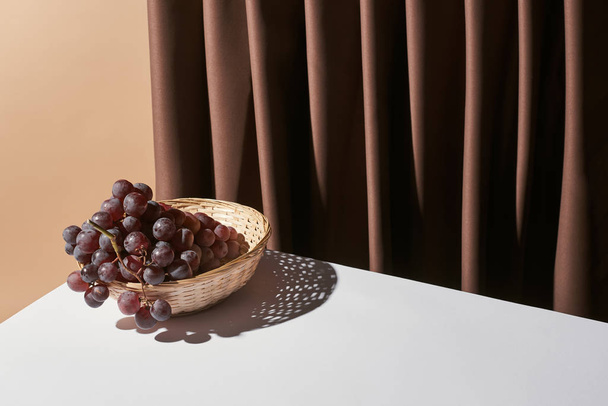 classic still life with grape in wicker basket on table near curtain isolated on beige - Photo, Image