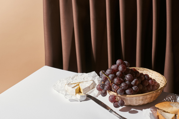 classic still life with grape in basket, brie cheese and baguette on table near curtain isolated on beige - Photo, Image